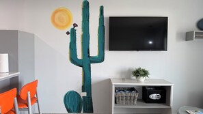 Une pièce décorée d’une grande œuvre d’art représentant un cactus, dotée d’un téléviseur à écran plat, d’une étagère blanche ornée d’une plante et de chaises orange.