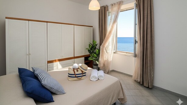 A bedroom with a bed, a bedside table with towels and a hat, a hanging light, a window with curtains, and a view of the sea. - Casa Baia Salata 2 (Imperia)