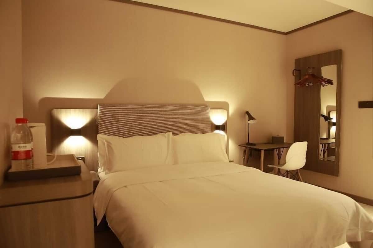 Quarto de hotel com cama, mesa de cabeceira, abajur, cadeira e espelho na parede.