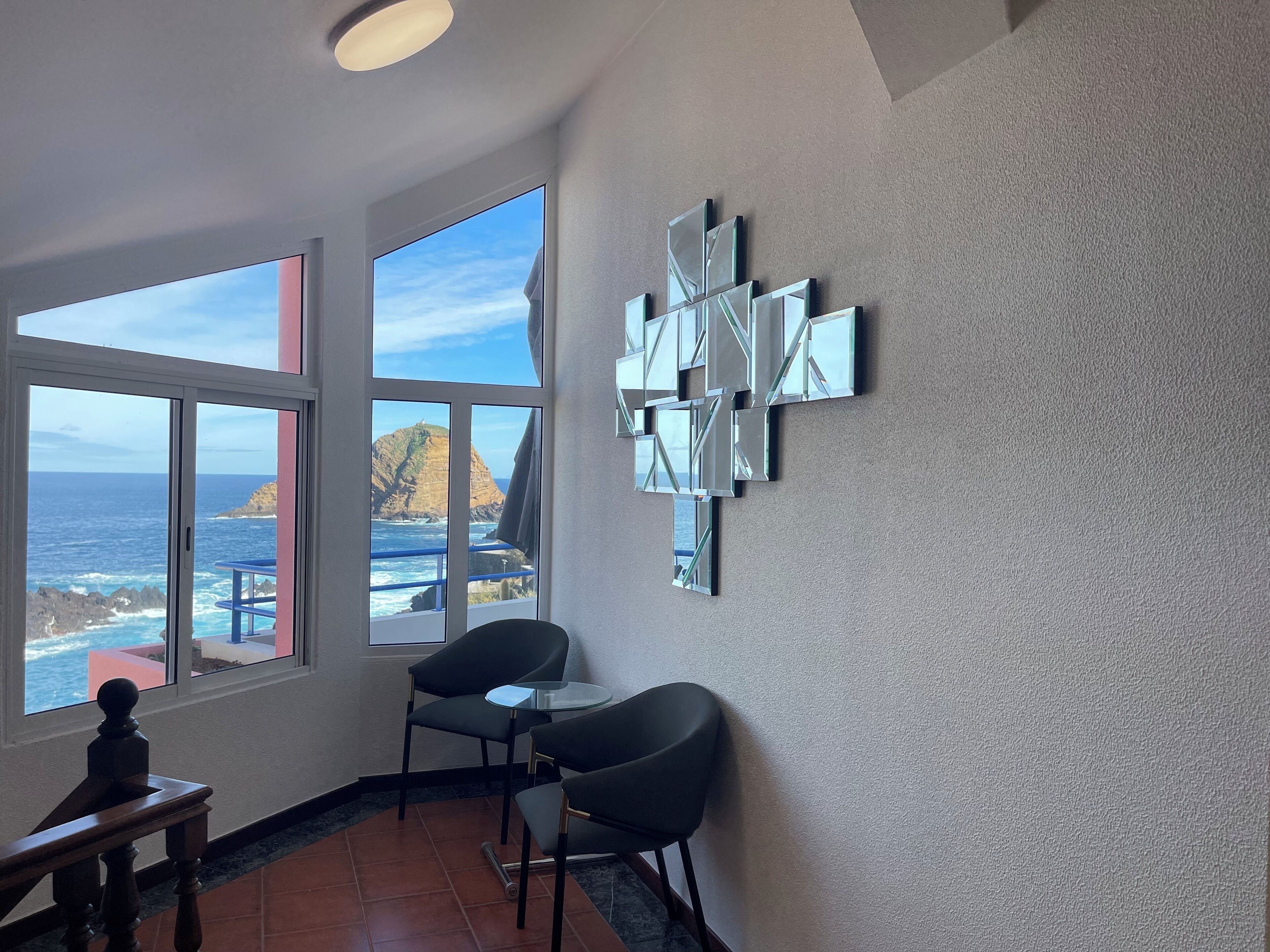 A room with large windows overlooking a coastal view, a modern wall art piece, and a small table with two chairs.