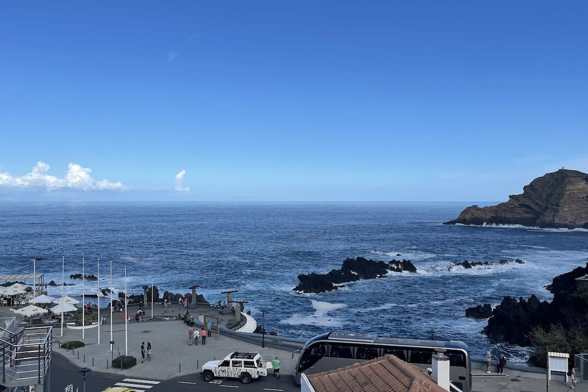 Vista costera con un promontorio rocoso, un paseo marítimo con asientos y sombrillas, y un cielo azul despejado.