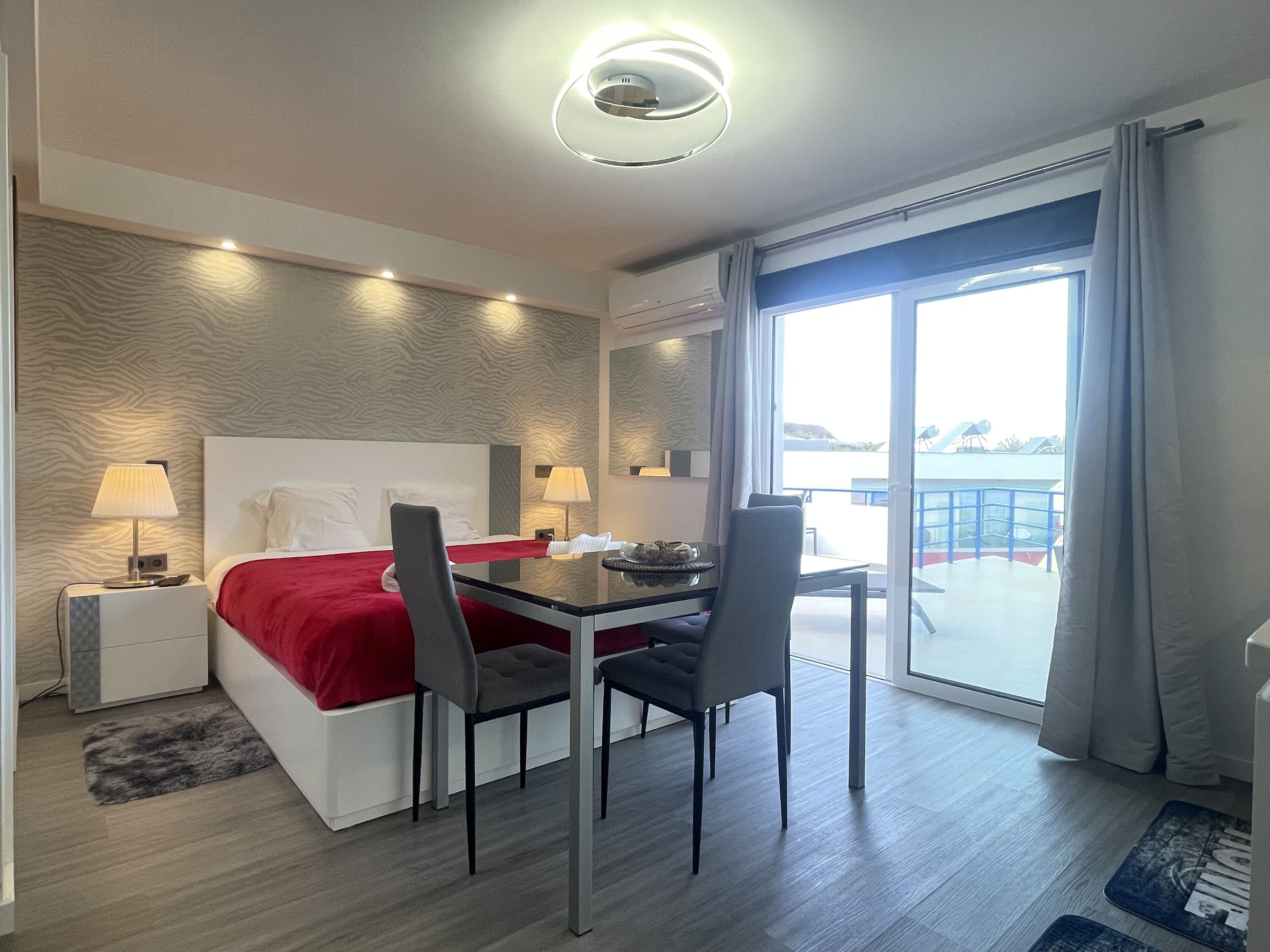 A hotel room with a bed, two bedside tables with lamps, a desk with chairs, and a view through a large window.