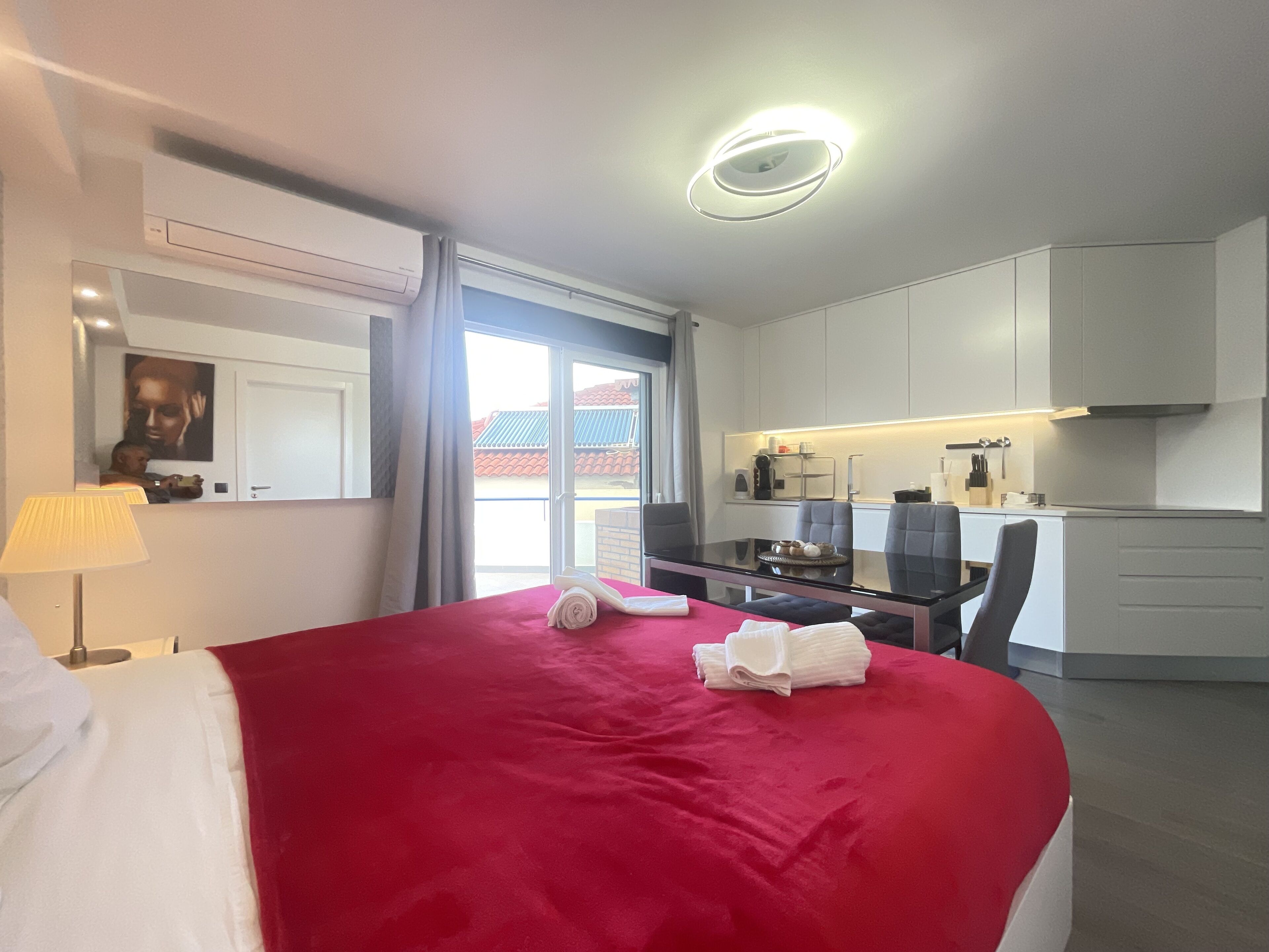 A hotel room with a large bed, a desk, a chair, a small kitchenette, and a window with a view of buildings.