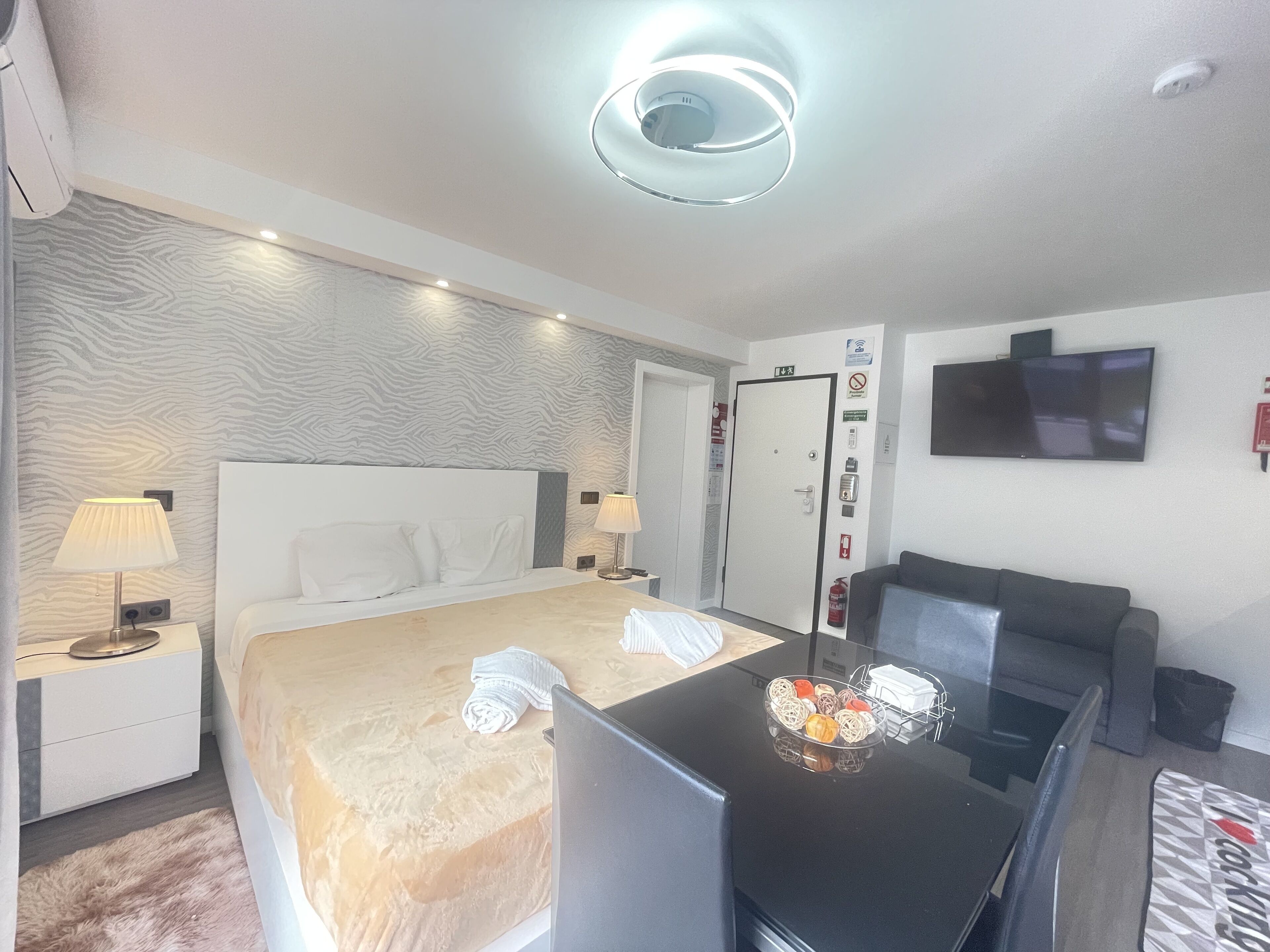 A modern hotel room with a large bed, bedside tables, a desk, a flat-screen TV, and a ceiling light fixture.