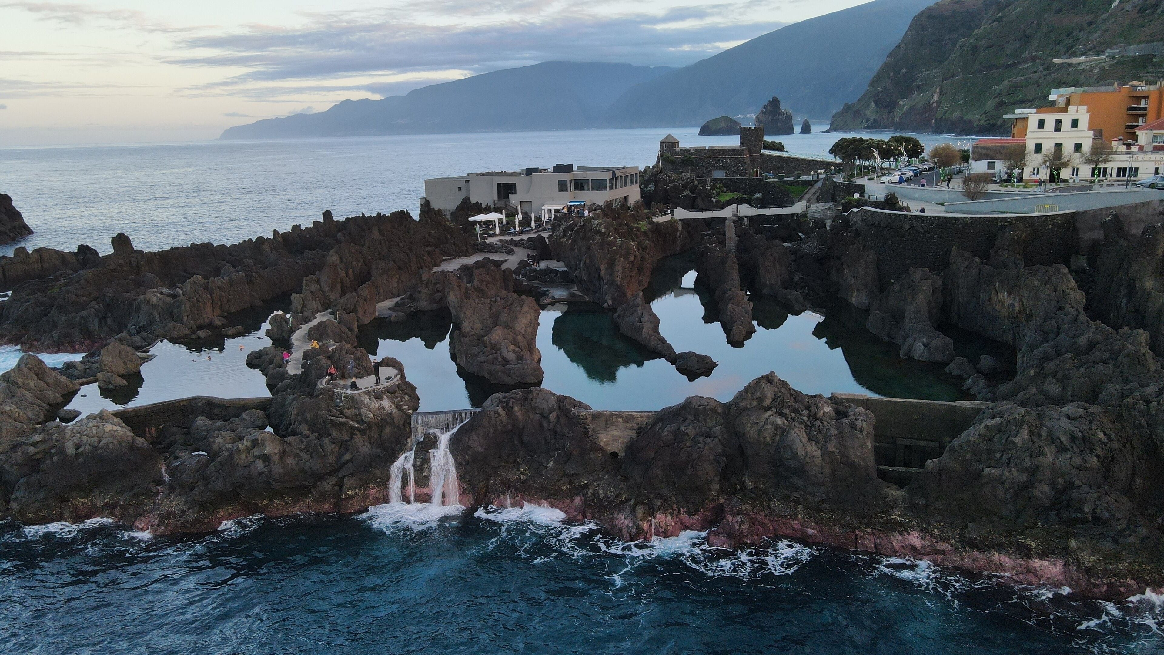Zona costera con formaciones rocosas, una cascada y edificios con balcones con vistas al mar.