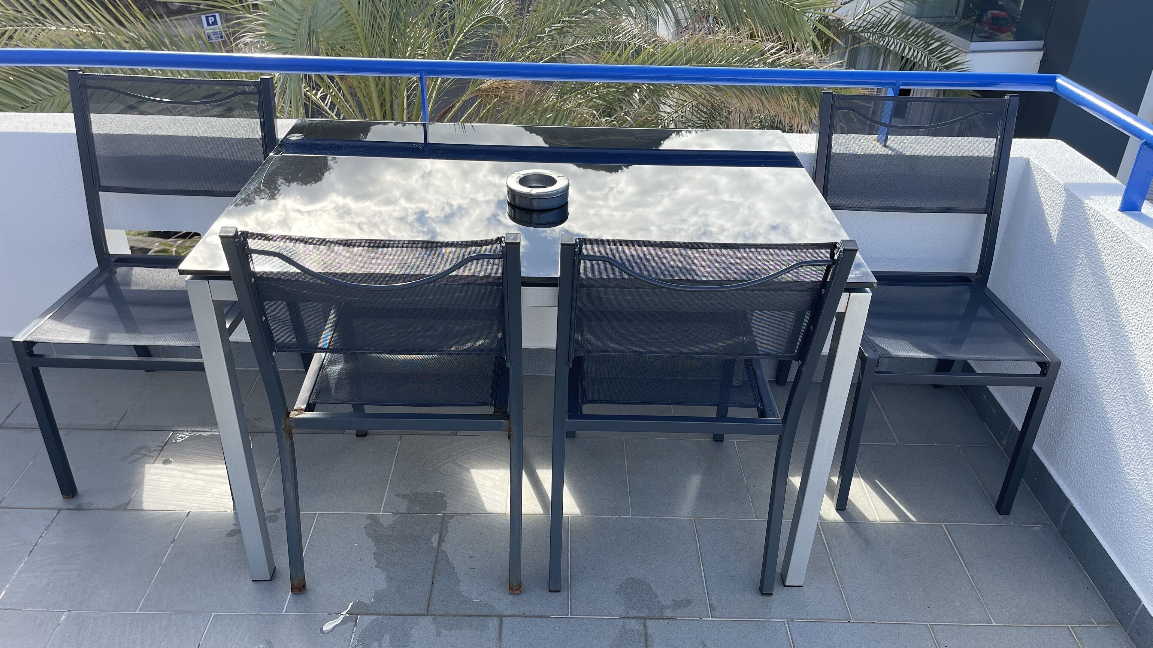 An outdoor patio with a table and chairs, a railing, and a view of palm trees.