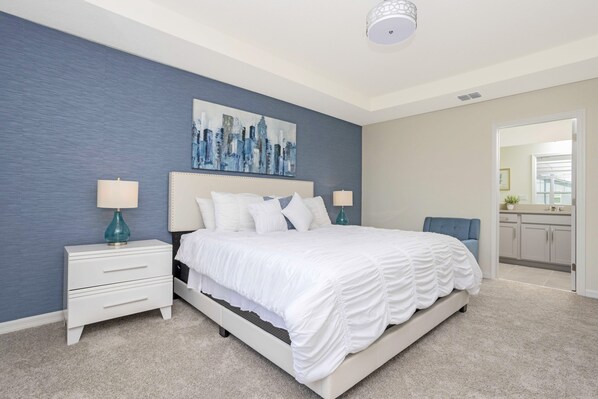 A bedroom with a large bed, two bedside tables, a cityscape painting, and a view of the kitchen through an open door.
