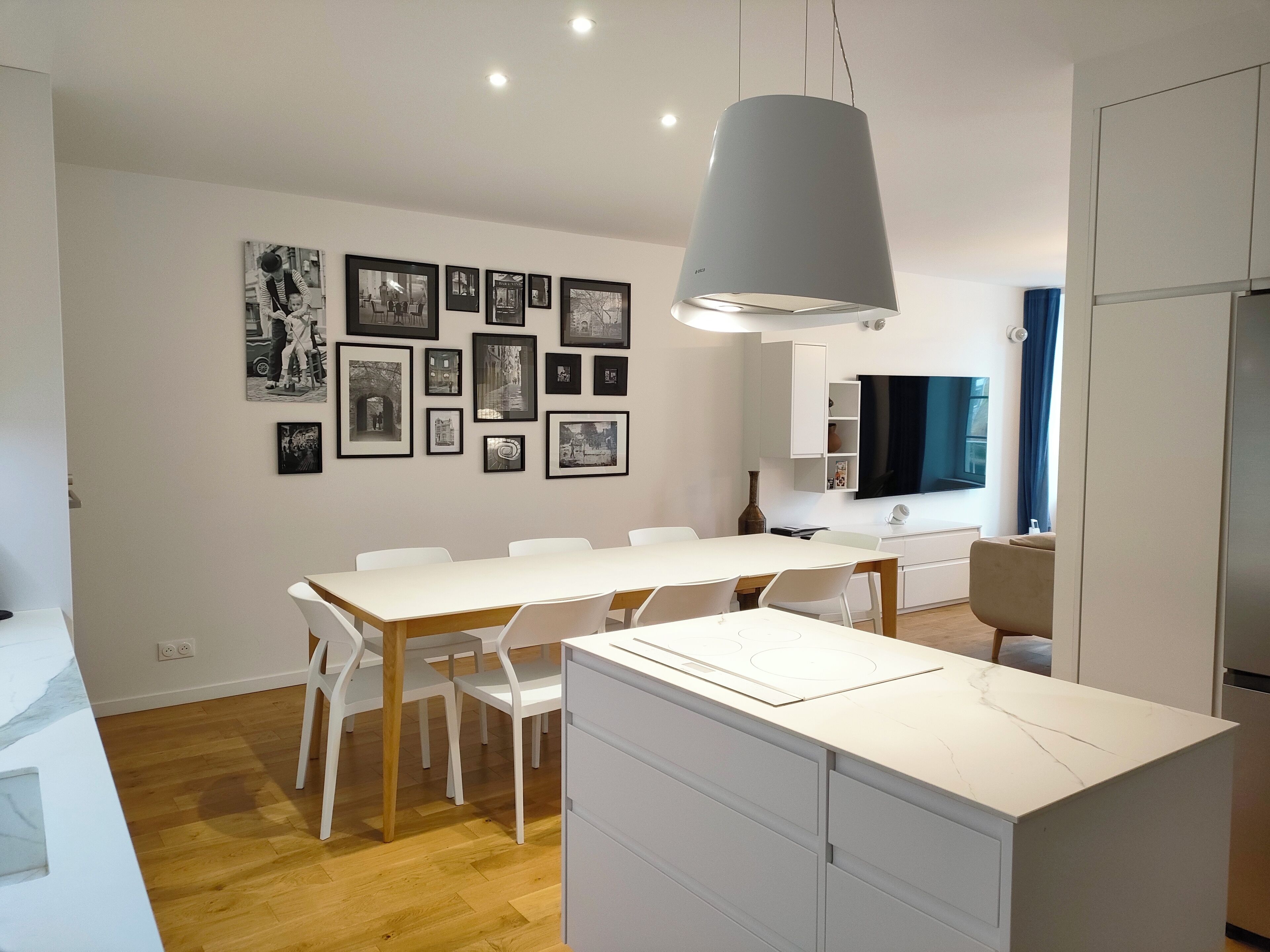 A modern kitchen with a white countertop, white chairs, and a dining table. There are framed pictures on the wall and a hanging light fixture.
