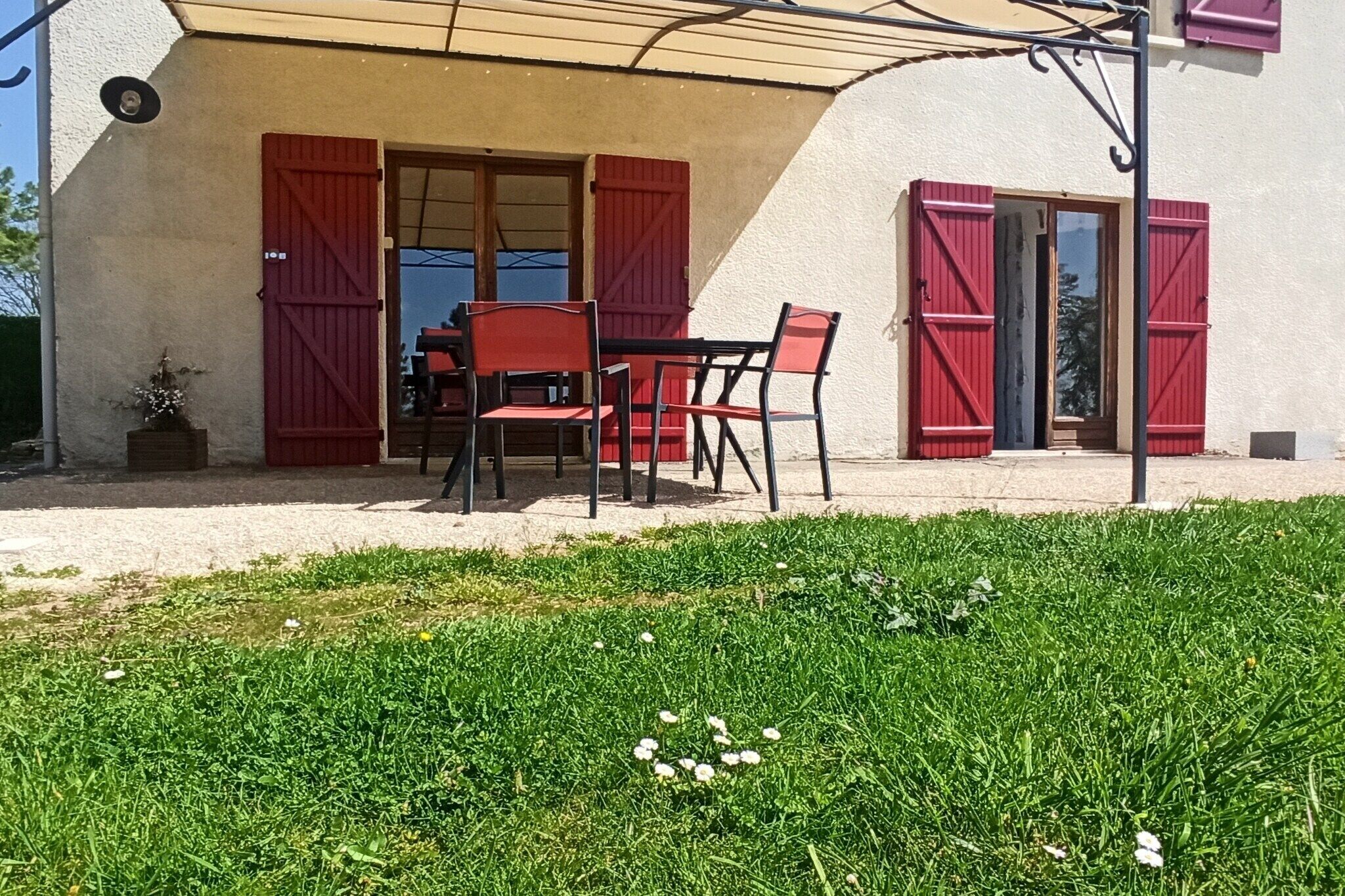 A patio with a table and chairs, a small garden area, and a building with red shutters.