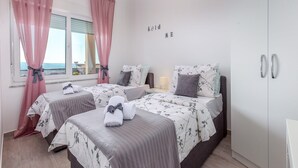 A bedroom with two beds, each with floral bedding and towels. There is a window with pink curtains, a bedside table with a lamp, and a wardrobe with silver handles. - Apartment with swimming pool near sea (Dramalj)