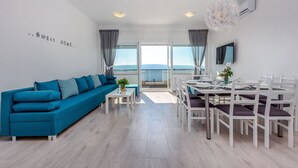 A modern living room with a blue sofa, dining table, and chairs. There is a large window with a view of the sea. - Apartment with swimming pool near sea (Dramalj)