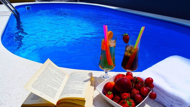 Escena junto a la piscina con un libro, dos bebidas y un cuenco de fruta.