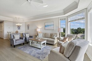 A spacious living room with a high ceiling, a ceiling fan, and large windows offering a view of the outdoors.