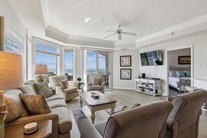 A spacious living room with a ceiling fan, large windows, and a view of the outdoors.