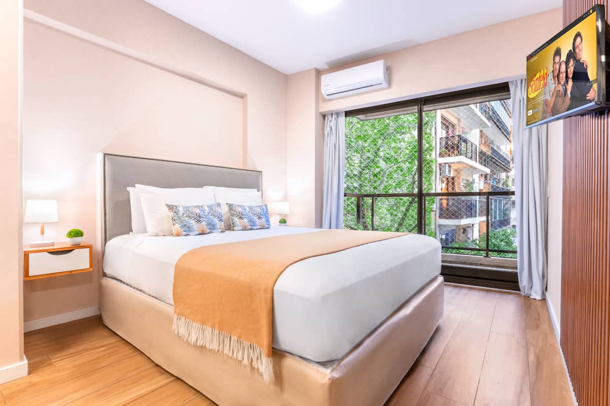 Quarto moderno com cama grande, mesa de cabeceira, aparelho de ar condicionado e janela com vista para edifícios e árvores.