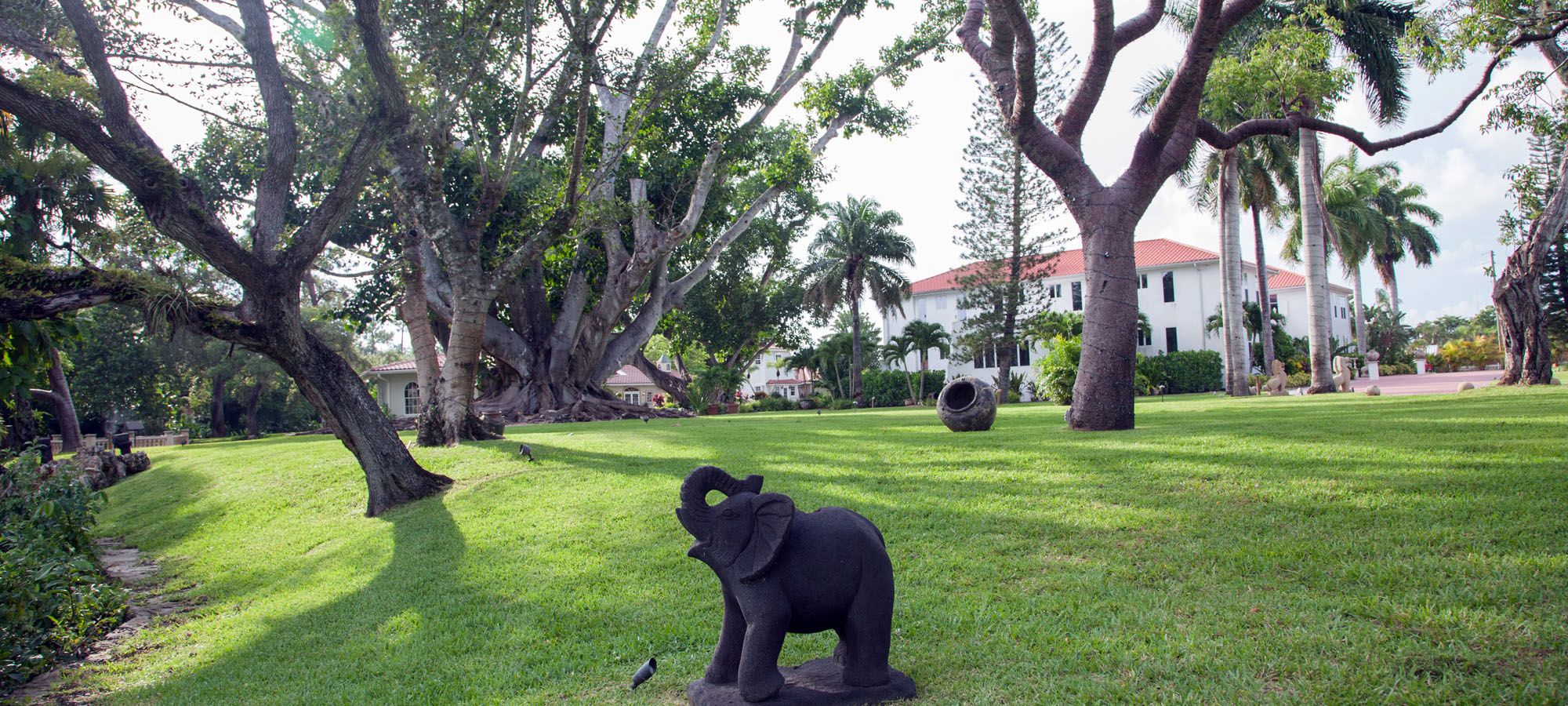 A grassy area with a large elephant sculpture, trees, and a building with a red roof in the background.