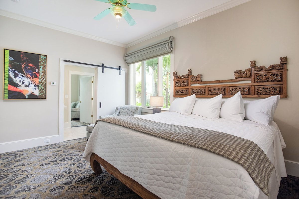 A bedroom with a large bed, a ceiling fan, a wooden headboard, and a painting on the wall.