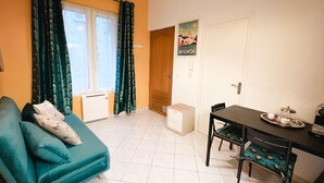 A living room with a teal sofa, a dining table with chairs, a small table with a teapot and cups, a window with curtains, and a door with a poster. - Charming apartment in the city center (Avignon)