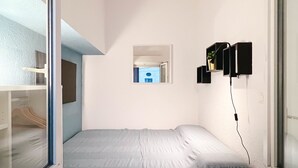 A bedroom with a bed, a window, a wall-mounted shelf, and a small window with a view of a blue building. - Charming apartment in the city center (Avignon)