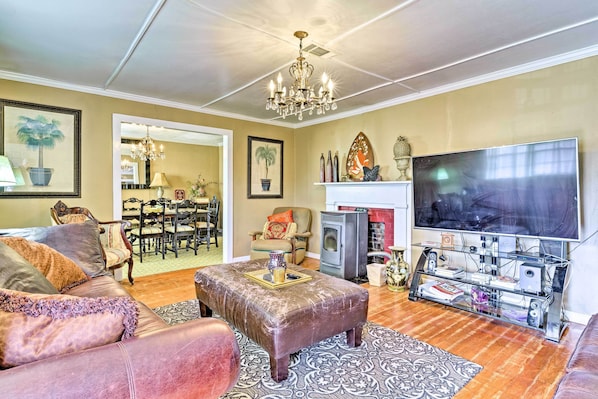 A living room with a sofa, ottoman, chandelier, flat-screen TV, fireplace, and a dining area with chairs and a table.