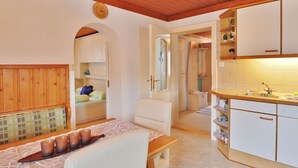 A compact room with a wooden ceiling, a bed with a headboard, a small table with cups, a kitchenette with white cabinets, and a bathroom visible through an open door. - Apartment in Niedernsill near Ski Slopes (Niedernsill)