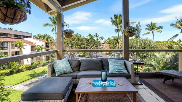 A covered outdoor seating area with a sofa, ottoman, and table, overlooking a lush garden and palm trees. - KKSR#8301 AMAZING 3 BEDR, TOP FLOOR CORNER UNIT: REMODELED, AIR CONDITIONING (Kailua Kona)