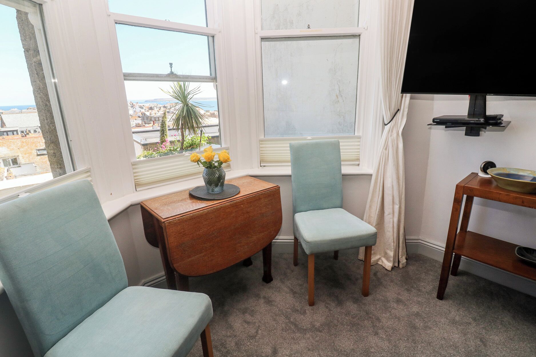 A room with two chairs, a small table with a vase of flowers, a television, and a view of buildings through the windows.
