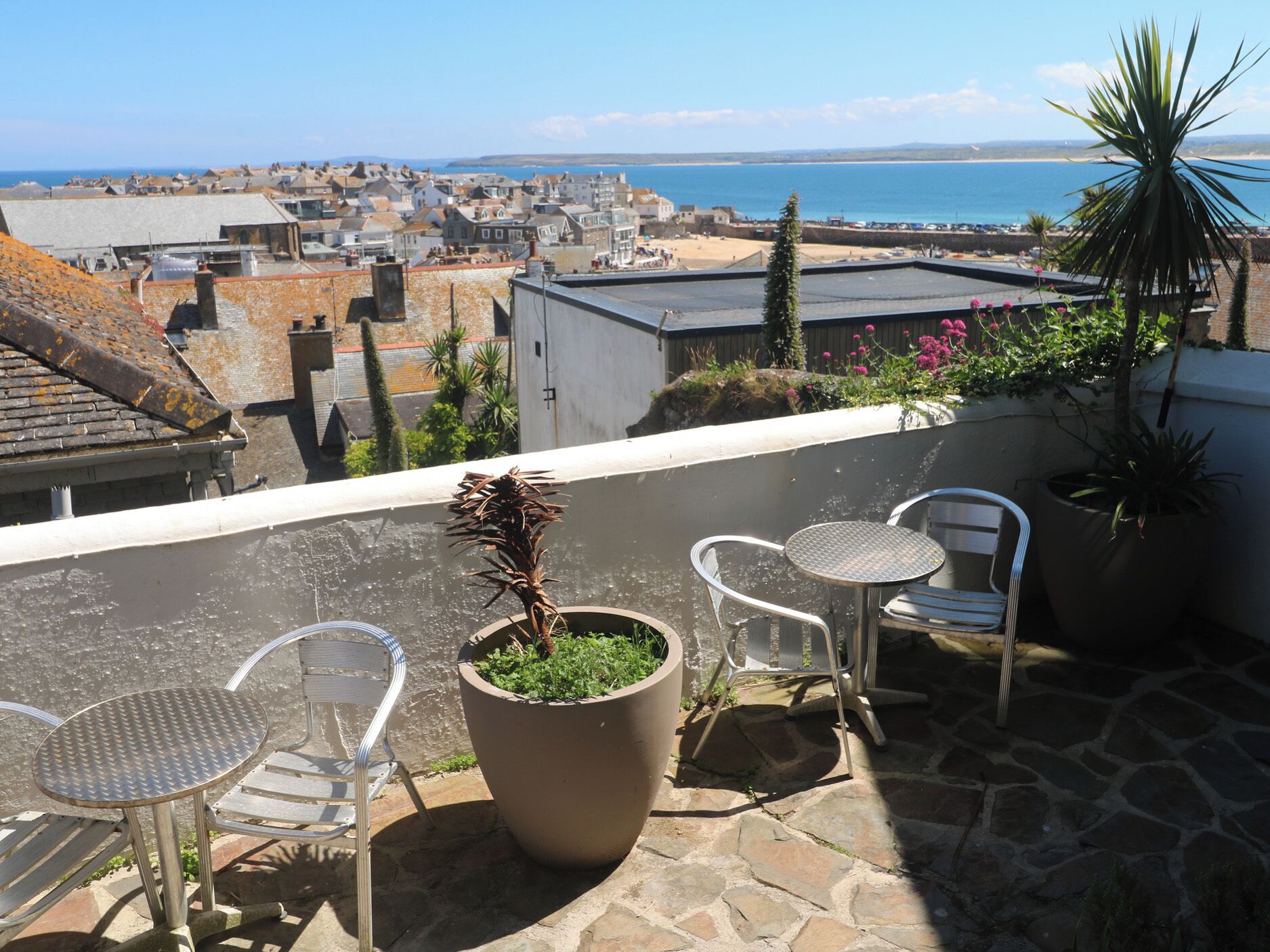 A rooftop terrace with a view of the sea, a potted plant, and two metal chairs.