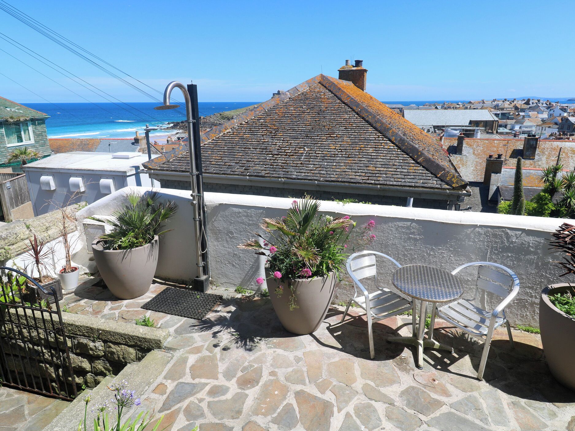 A rooftop terrace with potted plants, a metal chair, and a view of the ocean and town.