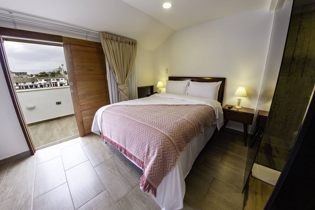 A hotel room with a bed, a nightstand, a lamp, and a view of the city through a balcony door.