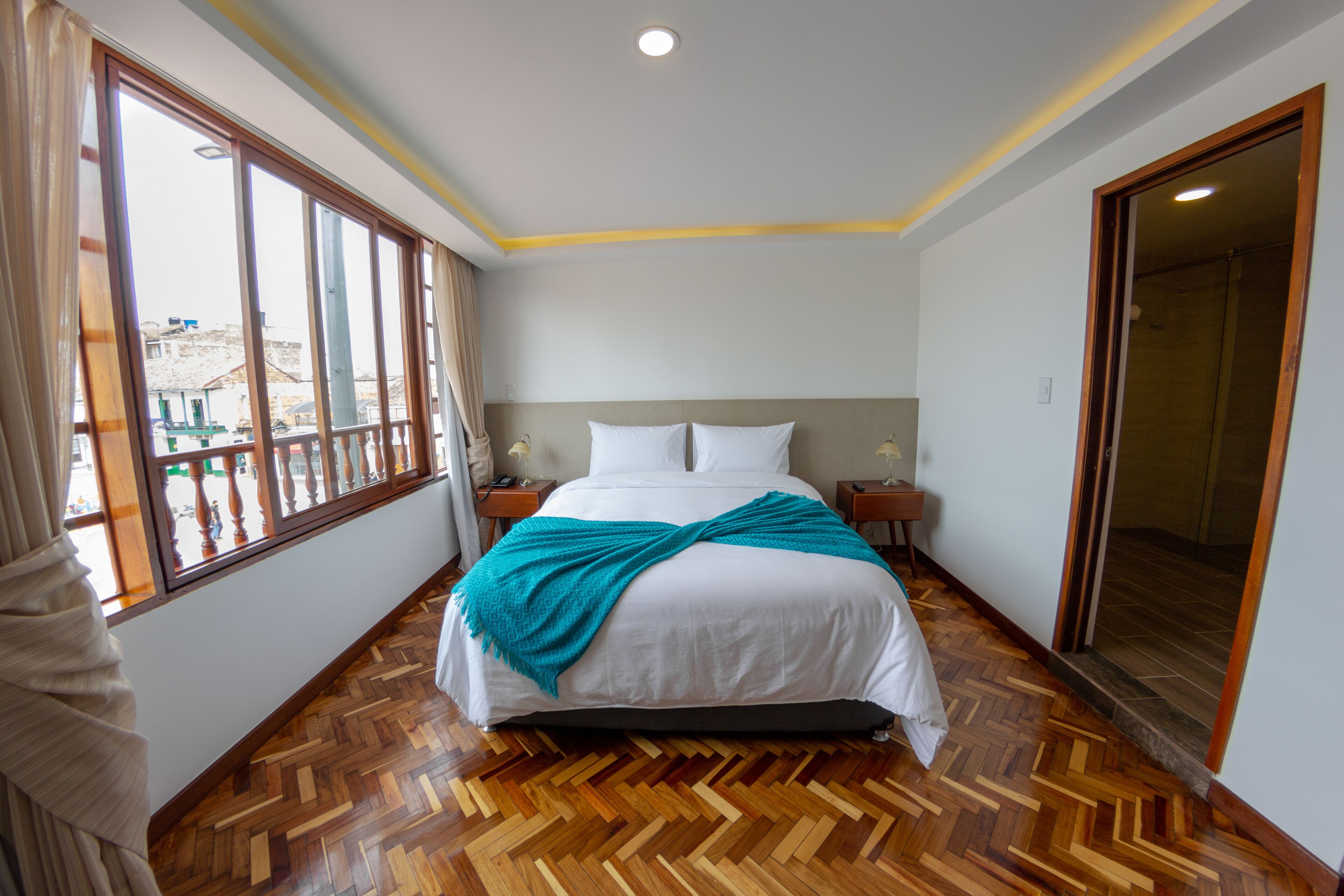 A hotel room with a large bed, wooden parquet flooring, a view through a window, and a door leading to a bathroom.