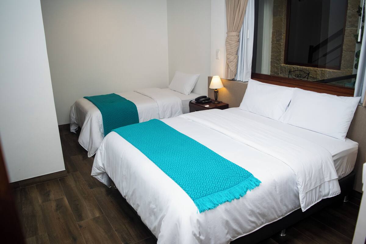 A hotel room with two beds, each with white bedding and a teal throw blanket. There is a nightstand with a lamp and a telephone. The room has wooden flooring and a large window with curtains.