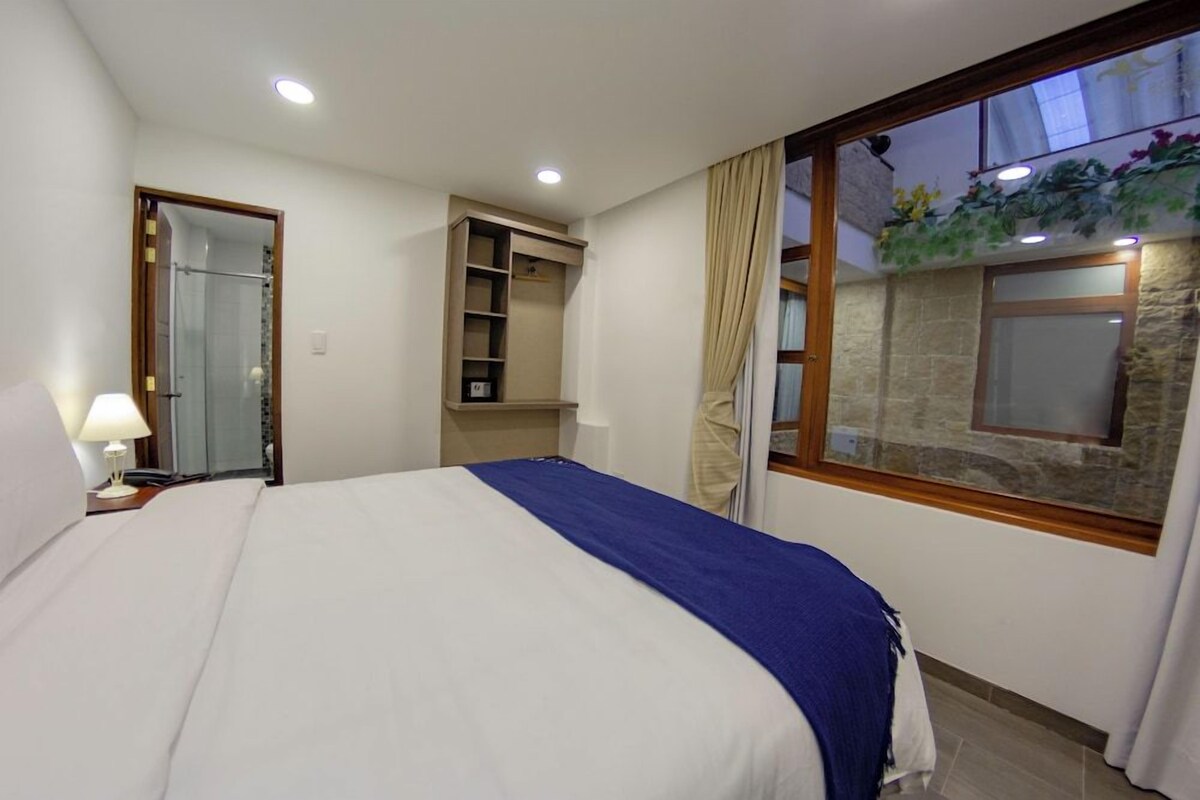 Camera d'albergo con un letto, un comodino, uno scaffale a parete e una finestra con tende, e una parete in pietra.