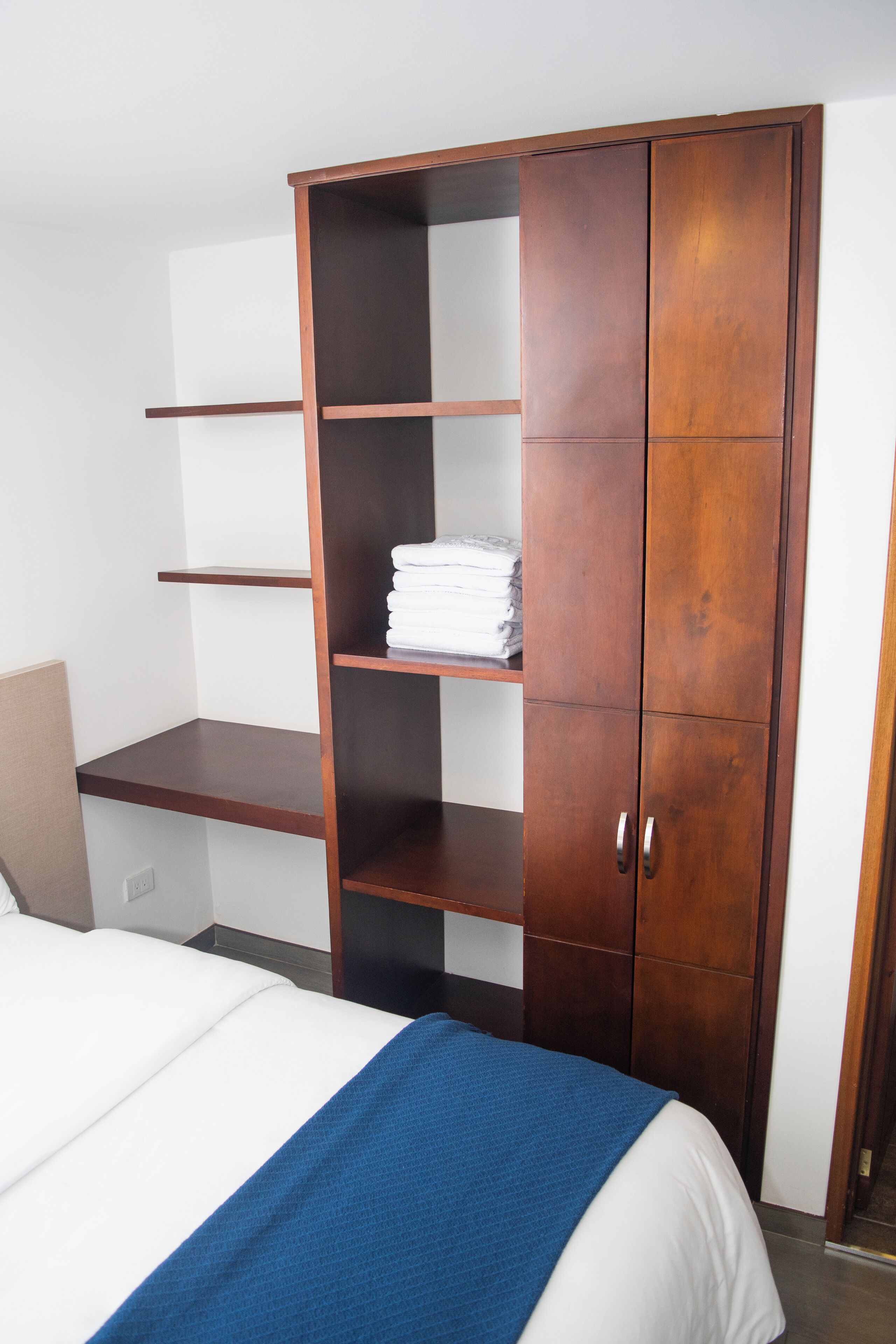 A bedroom with a bed, a wooden wardrobe with multiple shelves, and a white wall.