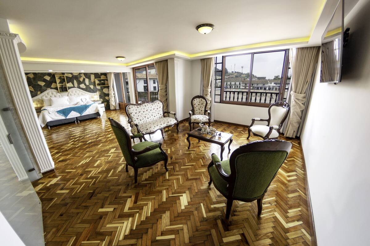A spacious living room with a herringbone wooden floor, a large bed with a geometric-patterned headboard, and a balcony with a view of the city.