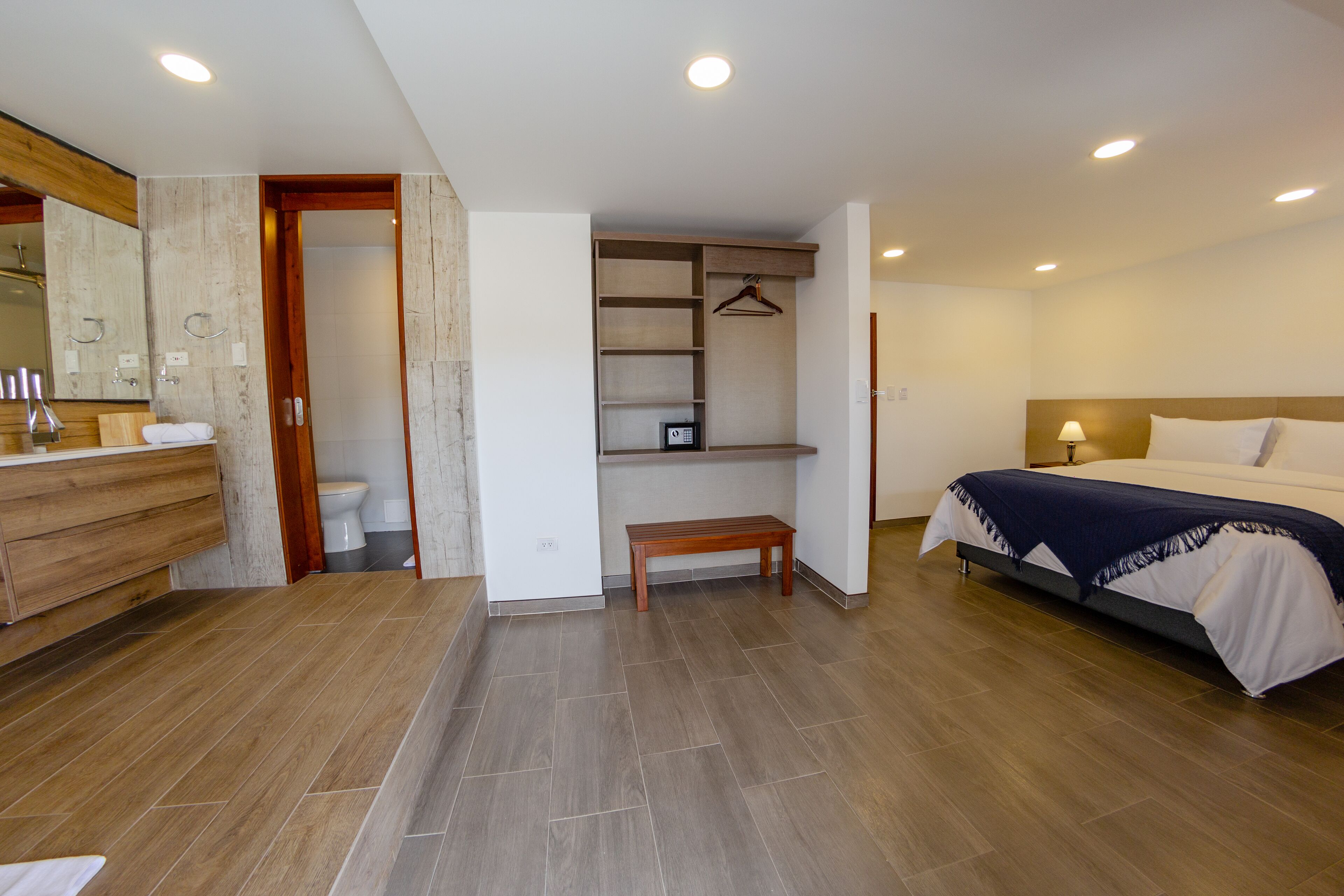 A modern bedroom with a wooden floor, a bed with a white and blue bedspread, a bedside table with a lamp, a wooden bench, and a built-in wardrobe with a hanging rack.