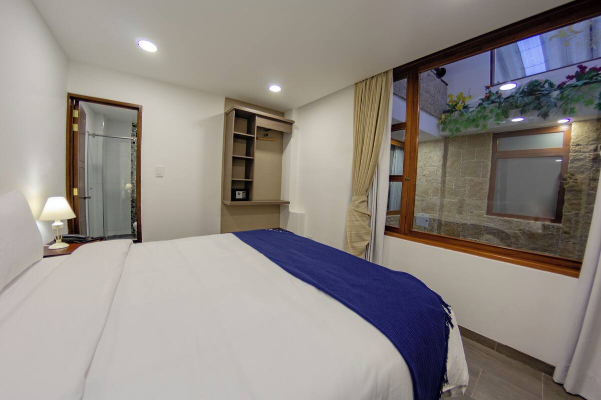 A bedroom with a bed, a nightstand, a built-in wardrobe, and a window with a view of a stone wall and greenery.