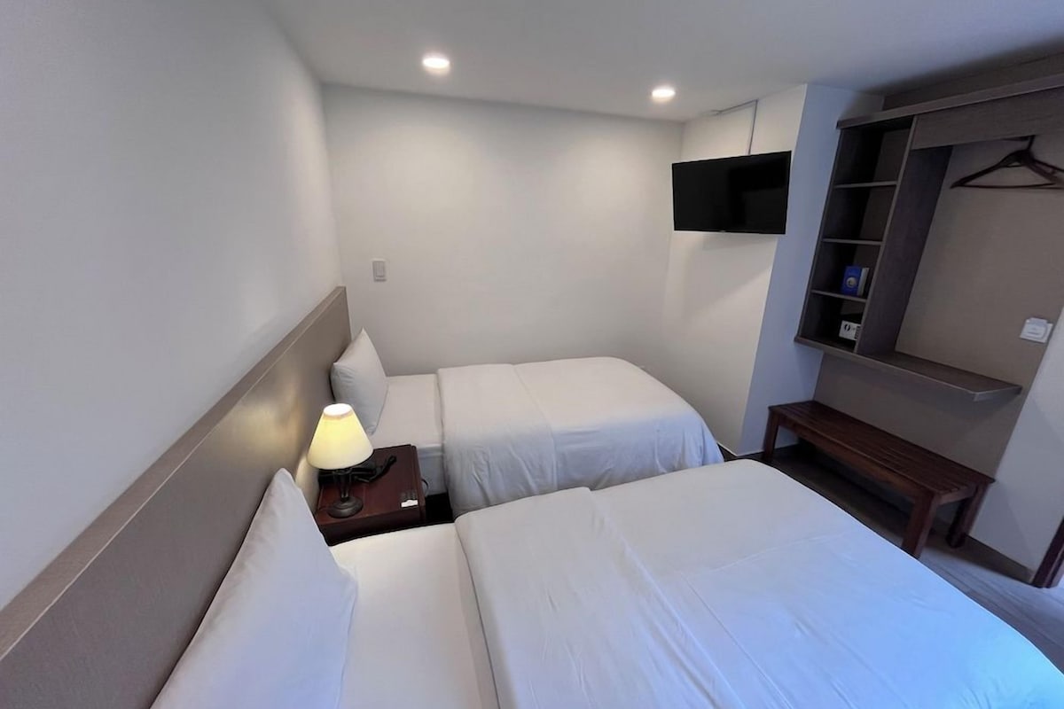 Camera d'albergo con due letti, un comodino con lampada, una TV a parete e un armadio con specchio.