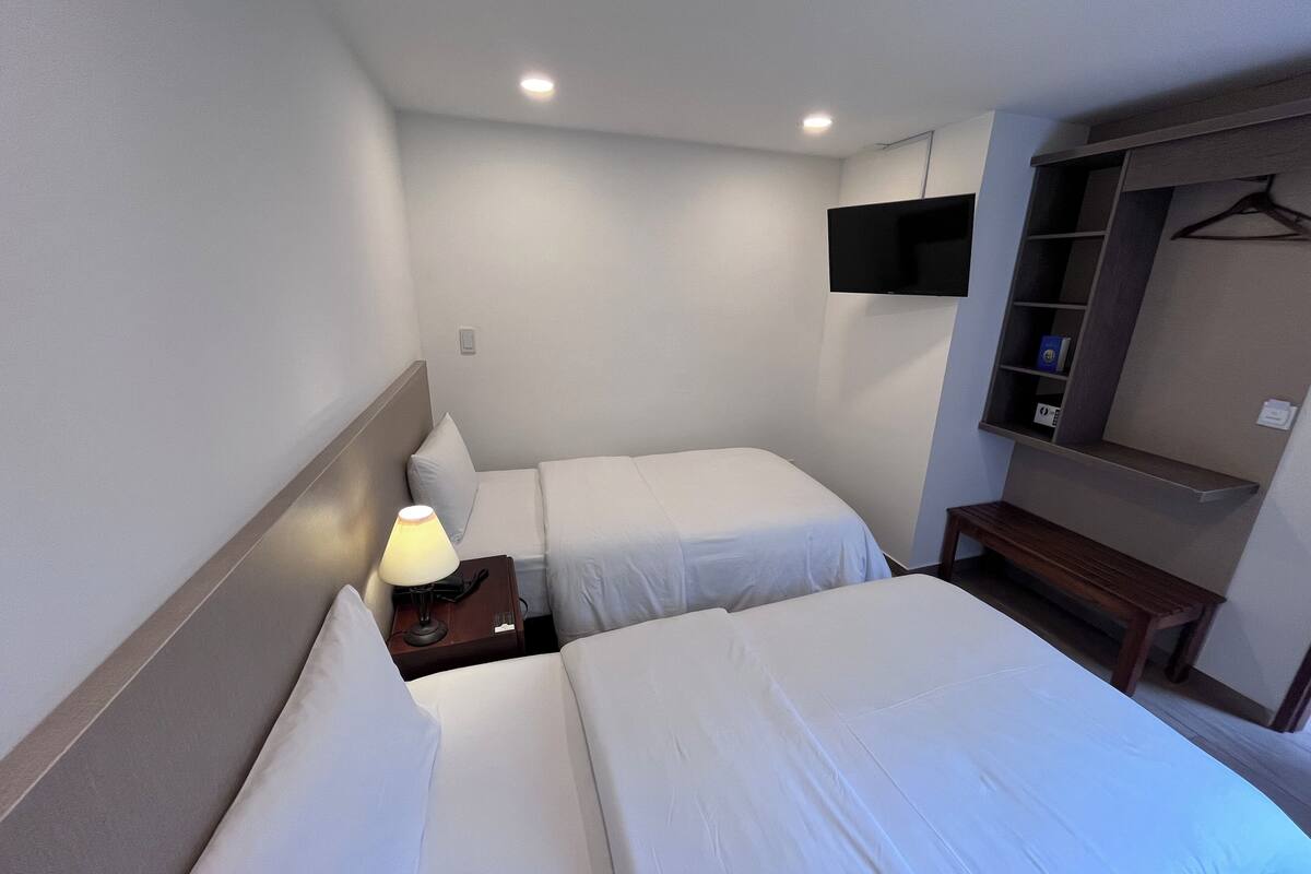 A hotel room with two beds, a bedside table with a lamp, a TV mounted on the wall, and a wardrobe with shelves.