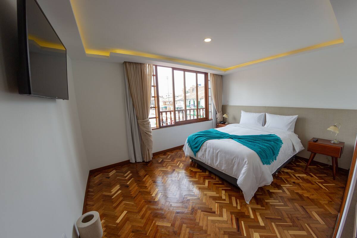 A bedroom with a large bed, wooden herringbone floor, a window with curtains, a bedside table with a lamp, and a flat-screen TV mounted on the wall.