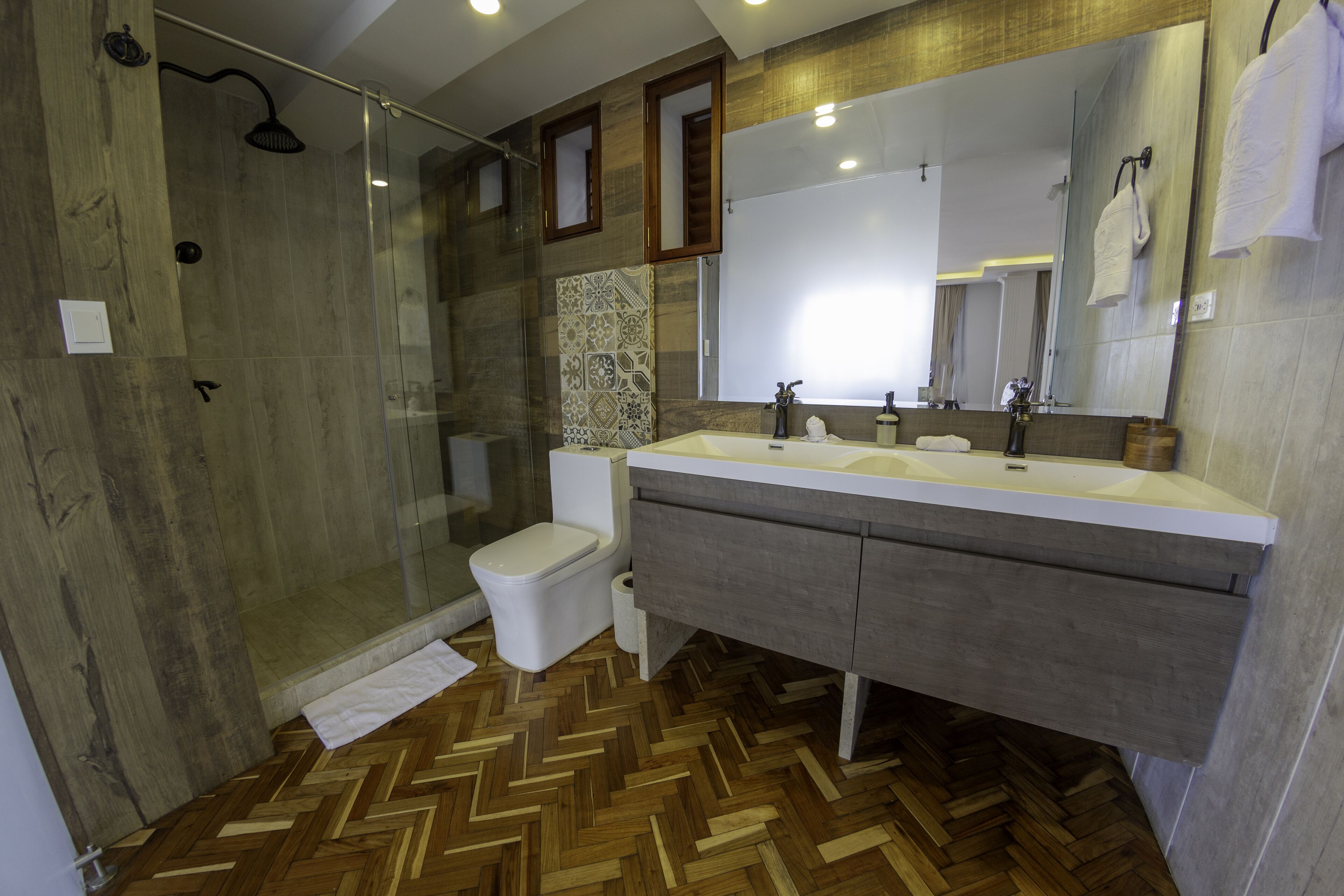 A modern bathroom with a herringbone wood floor, a glass shower enclosure, a white toilet, and a vanity with a large mirror.