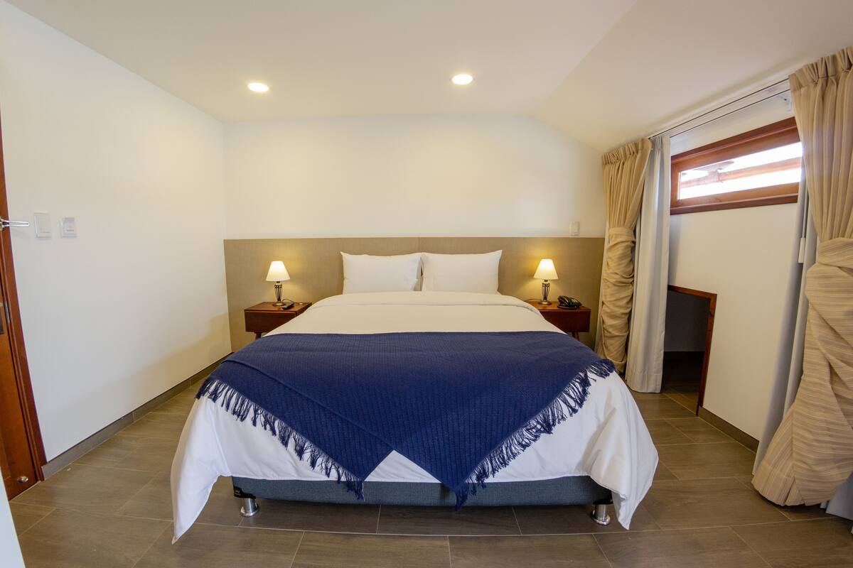 A hotel room with a bed, two bedside lamps, a wooden headboard, a window with curtains, and a door.