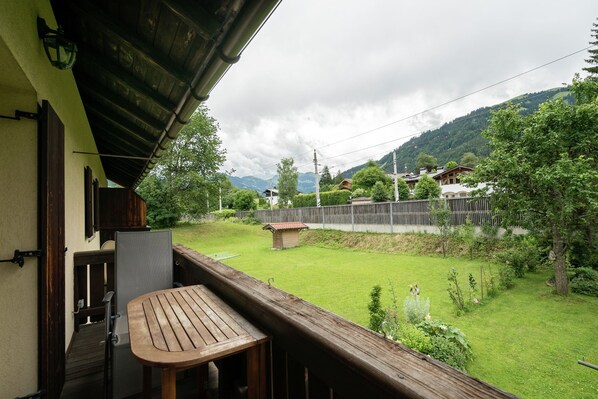 A balcony with a wooden table and chairs, overlooking a grassy yard with a wooden fence and houses in the background.