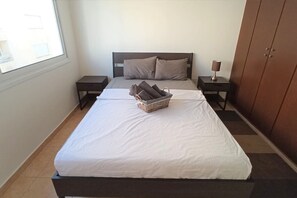 A neatly made bed with a white bedspread and pillows, a basket of folded towels, and a bedside lamp on a nightstand.