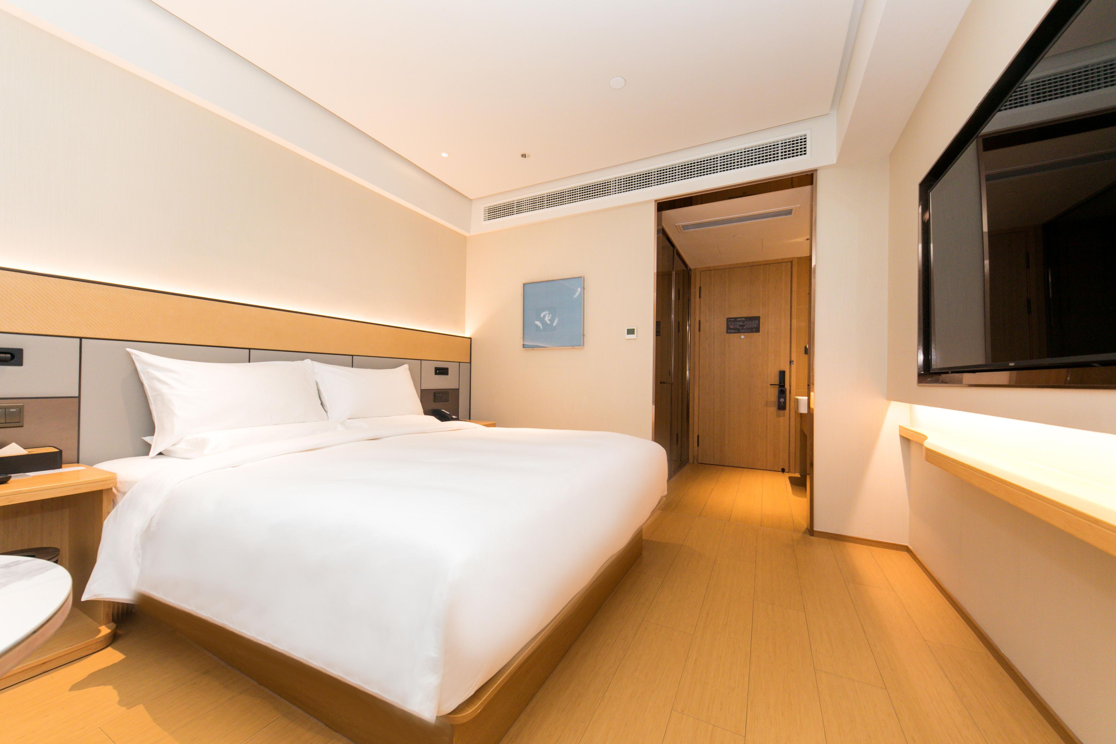 A hotel room with a large bed, bedside tables, a wooden headboard, a wall-mounted TV, and a door leading to another room.