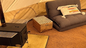 A cozy indoor camping setup with a tent, a portable stove, a checkered box, a grey sofa, and a patterned rug.