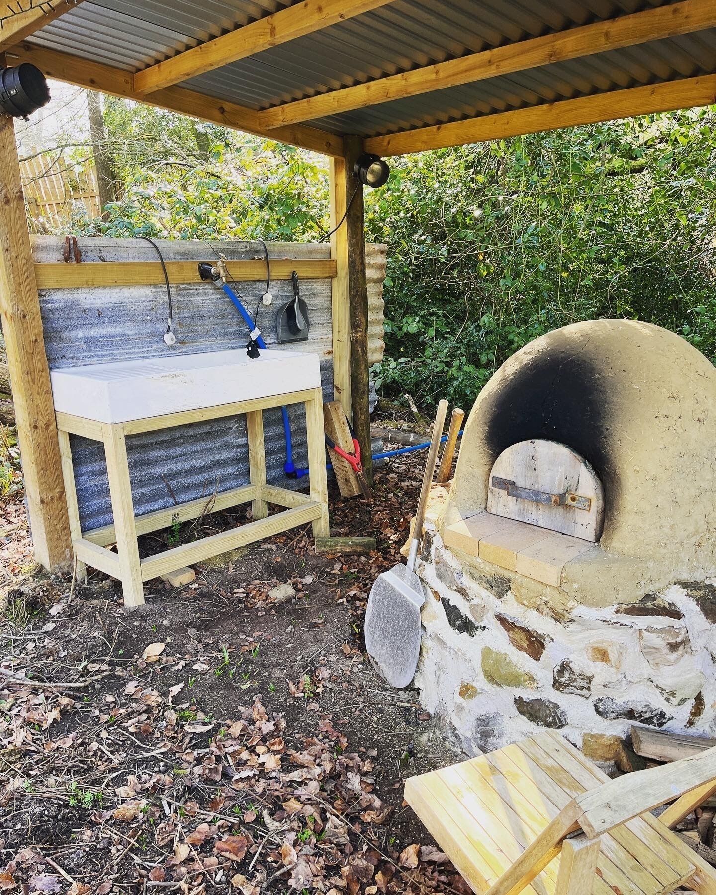 An outdoor cooking area with a stone oven, a wooden bench, and a metal grill.
