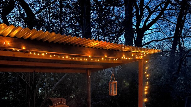 An outdoor stone oven with a fire, surrounded by a wooden pergola with string lights, a table with various items, and a folding chair.