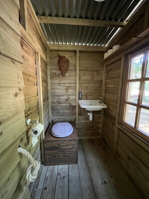 Un piccolo bagno in stile chalet, con pavimento e arredi in legno, una panca con il WC, un lavandino e una finestra.