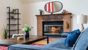 A living room with a brick fireplace, a black metal shelf with plants, a round mirror, and a blue sofa. - Forest 2BR chalet w/ hot tub & backyard (Gerrardstown)